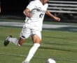 Player in a white soccer uniform dribbling a ball on a field during a night match, showcasing athletic skills.