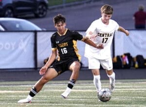 Two high school soccer players competing for the ball during a match, one in black and yellow, the other in white.