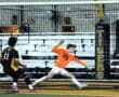 A soccer goalie in an orange jersey dives to block a shot on goal during a game, with a player in black nearby.