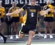 Soccer player in a black and yellow jersey shoots the ball during a night game, with cheerleaders in the background.