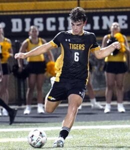 Soccer player in a black and yellow jersey shoots the ball during a night game, with cheerleaders in the background.