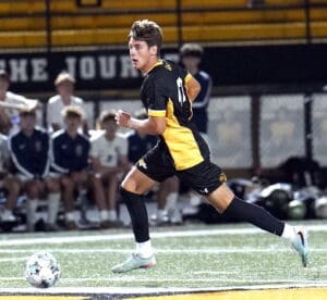 High school soccer player sprinting with the ball during a night game on the field, showcasing athletic skills.