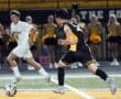 High school soccer match with players in action, one dribbling the ball while the other defends, cheerleaders in background.