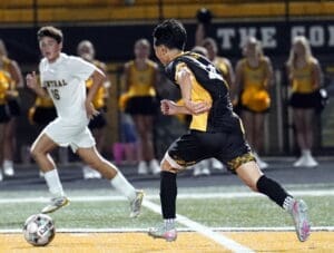 High school soccer match with players in action, one dribbling the ball while the other defends, cheerleaders in background.