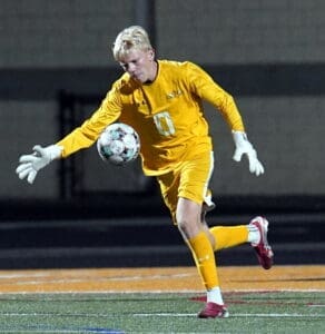 Goalkeeper in yellow kit catches soccer ball during night match on a field. Action shot showcasing athleticism and focus.