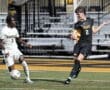 Two high school soccer players compete for the ball during a night game on a bright turf field.