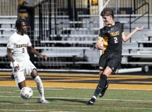Two high school soccer players compete for the ball during a night game on a bright turf field.