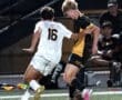 High school soccer players compete for possession of the ball during a nighttime match on a well-lit field.