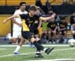 A soccer player kicks the ball during a game, with teammates and opponents in the background on the field.