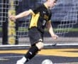 High school soccer player in black and yellow uniform kicking a ball on the field during a game.