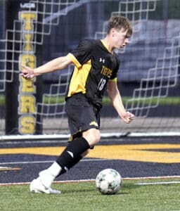 High school soccer player in black and yellow uniform kicking a ball on the field during a game.