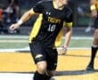 High school soccer player in black and yellow uniform dribbling a soccer ball on the field during a game.
