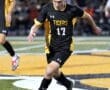 High school soccer player in black and yellow jersey dribbles the ball during a night game on a field.
