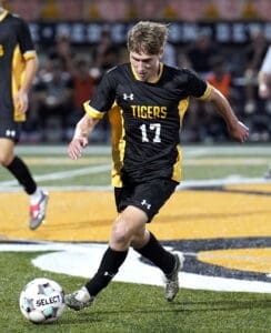 High school soccer player in black and yellow jersey dribbles the ball during a night game on a field.