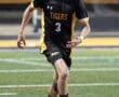 A soccer player in a black and gold uniform dribbles the ball on a field during a match, focus on athleticism and skill.