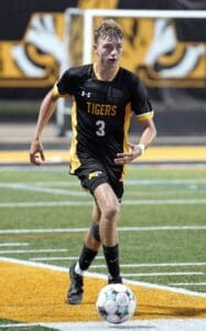 A soccer player in a black and gold uniform dribbles the ball on a field during a match, focus on athleticism and skill.