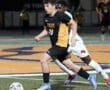 Young soccer player in a black and yellow uniform dribbles the ball while being pursued by an opponent on a field.