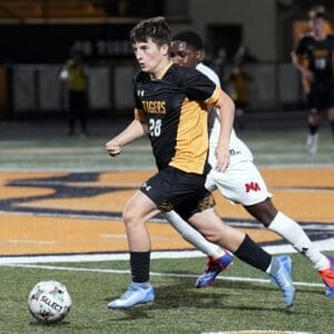 Young soccer player in a black and yellow uniform dribbles the ball while being pursued by an opponent on a field.