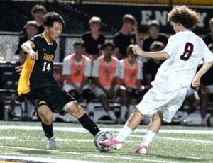 High school soccer match featuring players in action, one in black jersey and the other in white, with spectators in the background.