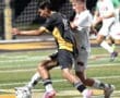 A soccer player in black battles for the ball against an opponent in white during a competitive match under bright lights.