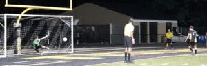 A soccer goalie dives to stop a penalty kick during a night match, with players and referees in the background.