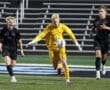 Goalkeeper in yellow chasing the ball as two opposing players celebrate during a night soccer match on the field.