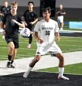 Two high school soccer players compete on the field, with one in white and the other in black, focused on the ball.