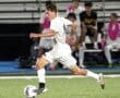 High school soccer player dribbling the ball during a night game, teammates cheering in the background on the sidelines.