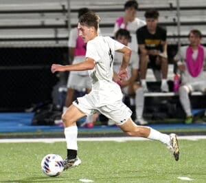 High school soccer player dribbling the ball during a night game, teammates cheering in the background on the sidelines.