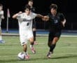 Two high school soccer players competing for the ball during a night game, one in white and one in black jerseys.
