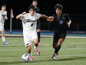 Two high school soccer players competing for the ball during a night game, one in white and one in black jerseys.