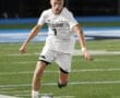 High school soccer player in white uniform dribbles the ball on a field during an evening match.
