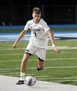 High school soccer player in white uniform dribbles the ball on a field during an evening match.