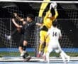 Goalkeeper in yellow leaps to catch the soccer ball amid players in black and white uniforms during a night match.