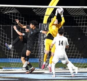 Goalkeeper in yellow leaps to catch the soccer ball amid players in black and white uniforms during a night match.