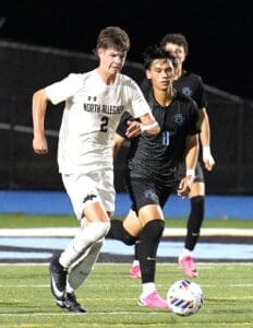North Allegheny soccer player sprinting with the ball while being pursued by an opponent on a lit field.