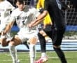 Two soccer players compete for the ball during a nighttime match, with a goalkeeper in the background.