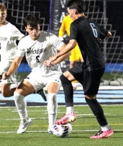 Two soccer players compete for the ball during a nighttime match, with a goalkeeper in the background.