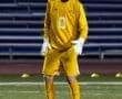 Goalkeeper in a yellow uniform stands ready on the field, focused on the soccer ball during a night game.