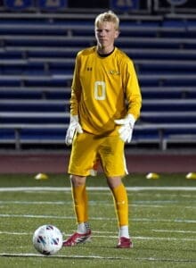 Goalkeeper in a yellow uniform stands ready on the field, focused on the soccer ball during a night game.