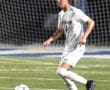 Player in a white soccer uniform dribbling a ball on a grassy field during a night game. Goal net in background.