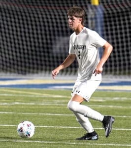 Player in a white soccer uniform dribbling a ball on a grassy field during a night game. Goal net in background.