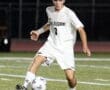 High school soccer player in white jersey dribbles a ball on a well-lit turf field during a night game.