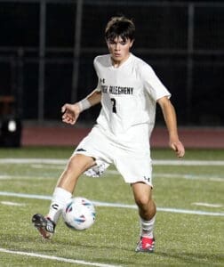 High school soccer player in white jersey dribbles a ball on a well-lit turf field during a night game.