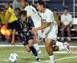 Two soccer players compete for the ball during a vibrant high school soccer match under bright stadium lights.