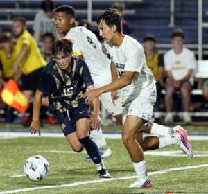 Two soccer players compete for the ball during a vibrant high school soccer match under bright stadium lights.