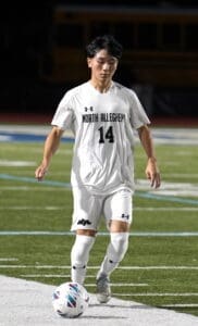 A soccer player in a white North Allegheny jersey dribbles a ball on a field at night.