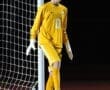 Goalkeeper in yellow uniform ready to defend during a night soccer match with a ball at his feet.