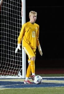 Goalkeeper in yellow uniform ready to defend during a night soccer match with a ball at his feet.