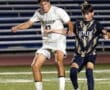 Two high school soccer players compete for the ball on a well-lit field during an evening match.